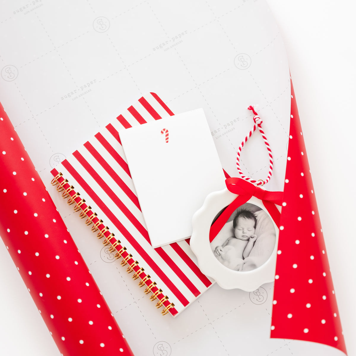Red and white holiday-themed gift set featuring a red and white striped spiral notebook, a blank white notepad with a candy cane icon, a red ribbon, and a scalloped photo ornament with a red ribbon