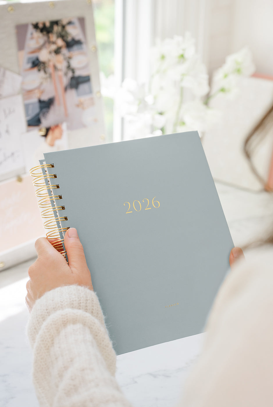 Person holding a blue planner with '2026' on it, sitting in a bright room.
