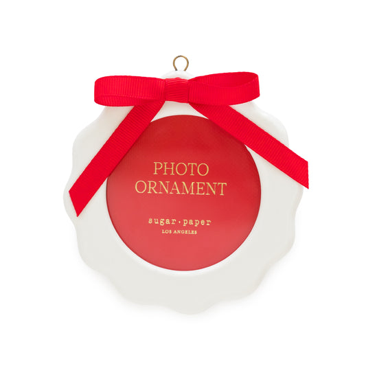 A white scalloped Christmas ornament with a red circular photo opening center, a red ribbon bow, and gold lettering that reads 'PHOTO ORNAMENT' and 'sugar paper LOS ANGELES'.