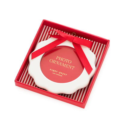 A white scalloped-edge photo ornament with a red ribbon bow, presented inside a red gift box with red-and-white striped interior. The ornament has a red center with gold text reading 'PHOTO ORNAMENT' and smaller 'sugar paper' branding.