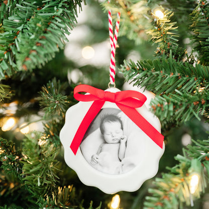 A white scalloped ceramic Christmas ornament hanging on a green Christmas tree with warm lights. The ornament features a printed black-and-white photo of a sleeping newborn baby and is tied with a bright red ribbon bow and red-and-white striped hanging string.