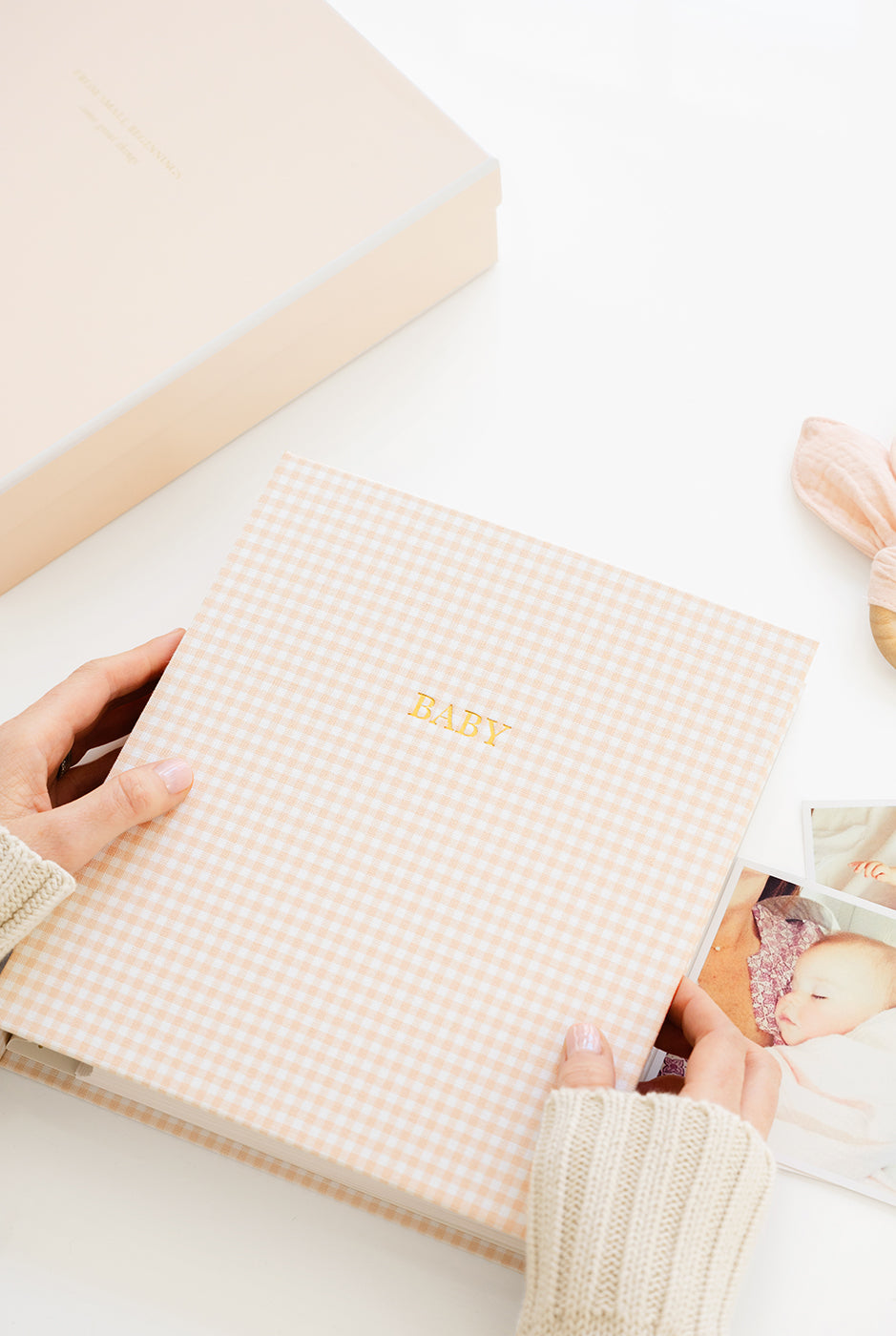 Person holding a pink gingham baby book with 'Baby' printed on it, on a white background.