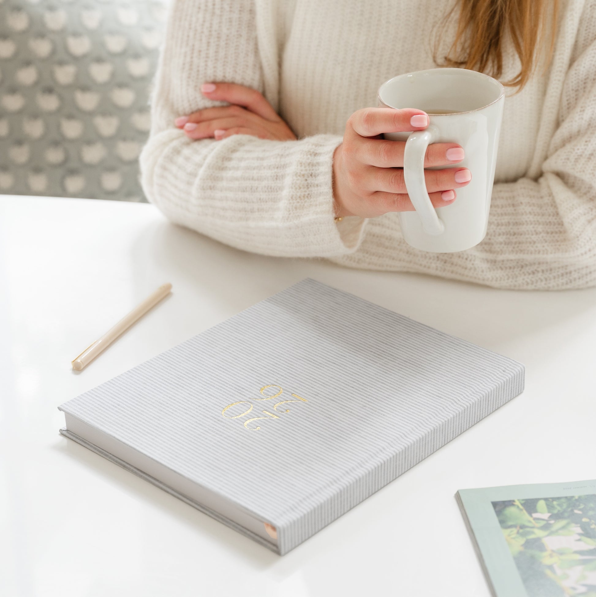 Person holding a white mug with a 2026 grey stripe fabric planner on the table in front of her
