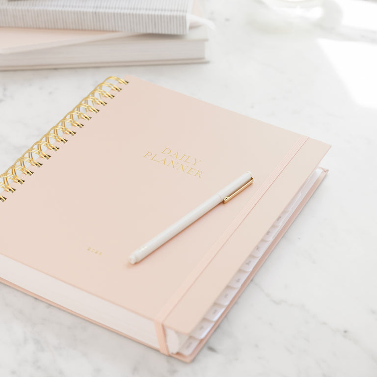 Blush Pink spiral planner with gold spiral binding and a white pen on a marble surface