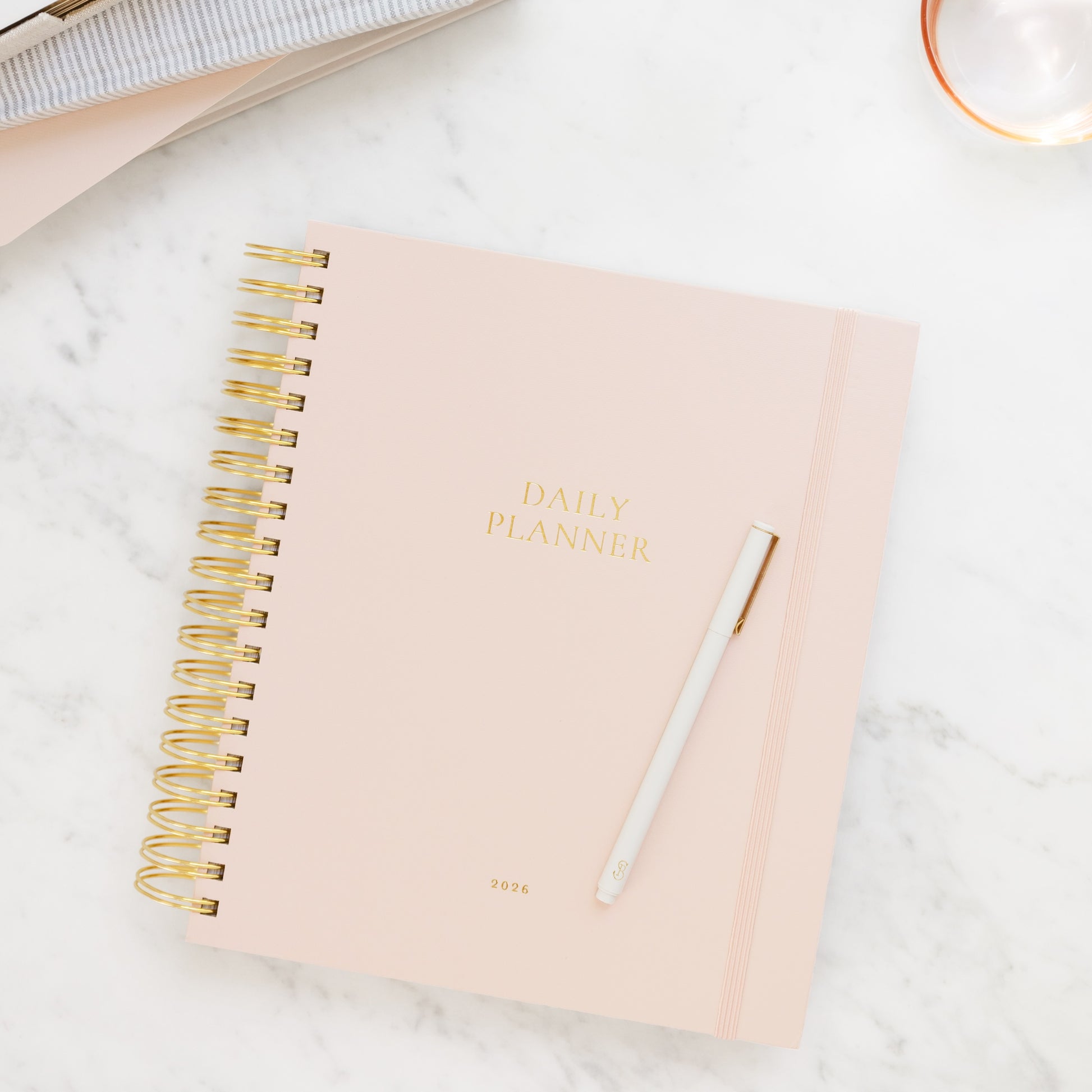 Blush pink daily spiral planner with gold spiral binding on a marble surface with journals in background