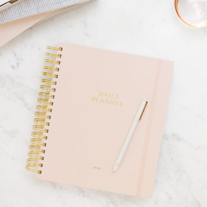 Blush pink daily spiral planner with gold spiral binding on a marble surface with journals in background