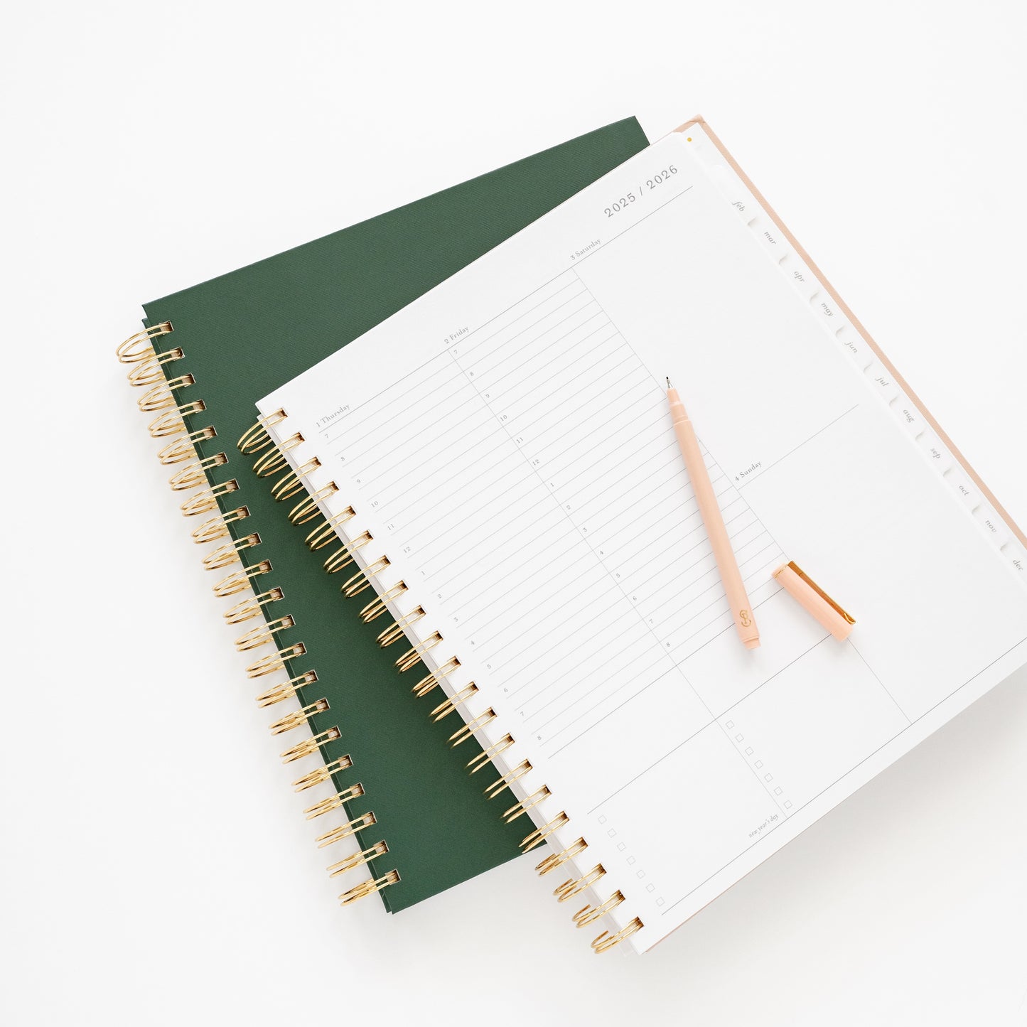 Two spiral-bound planners, one green and one open to weekly page, with a pink pen on top