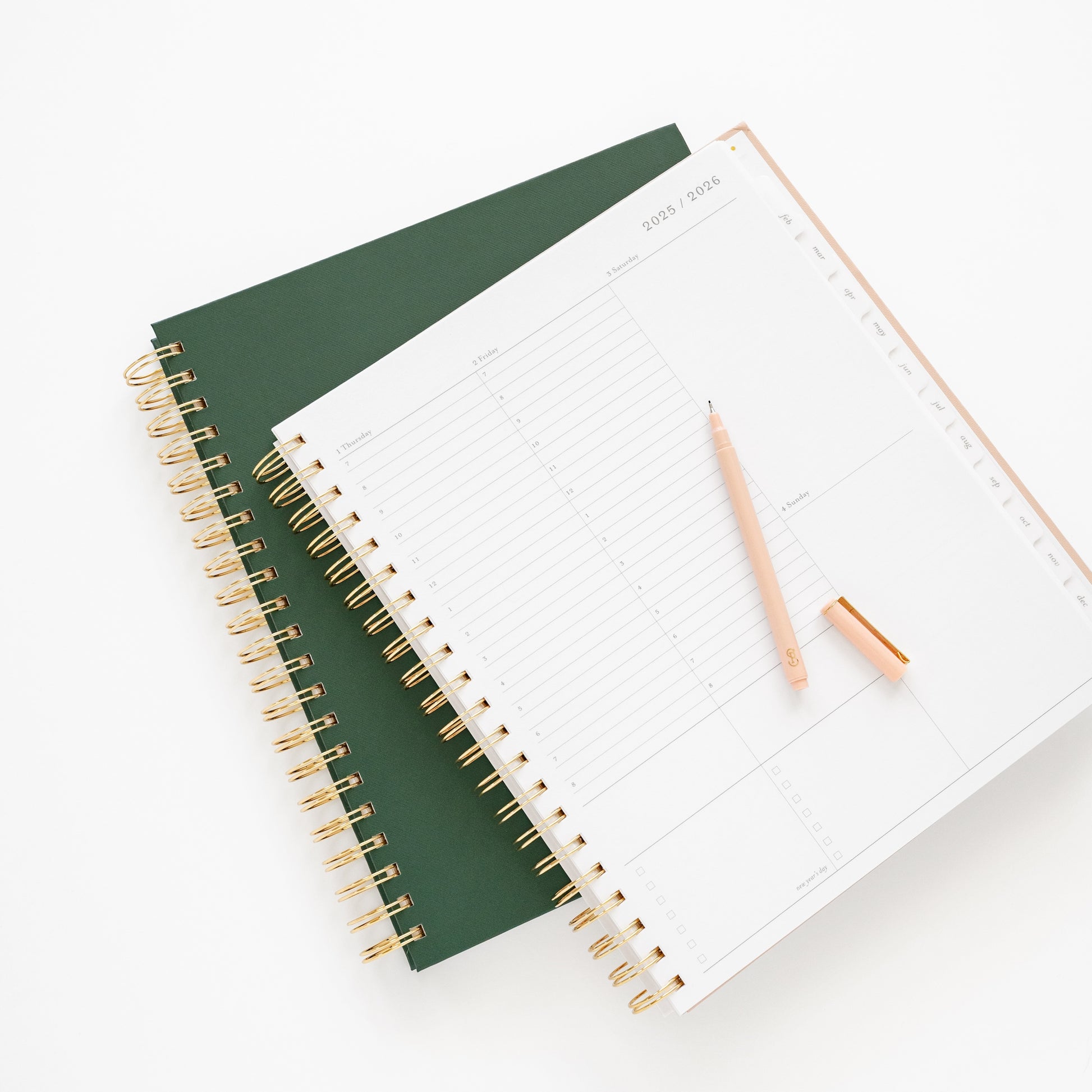 Two spiral-bound planners, one green and one open to weekly page, with a pink pen on top