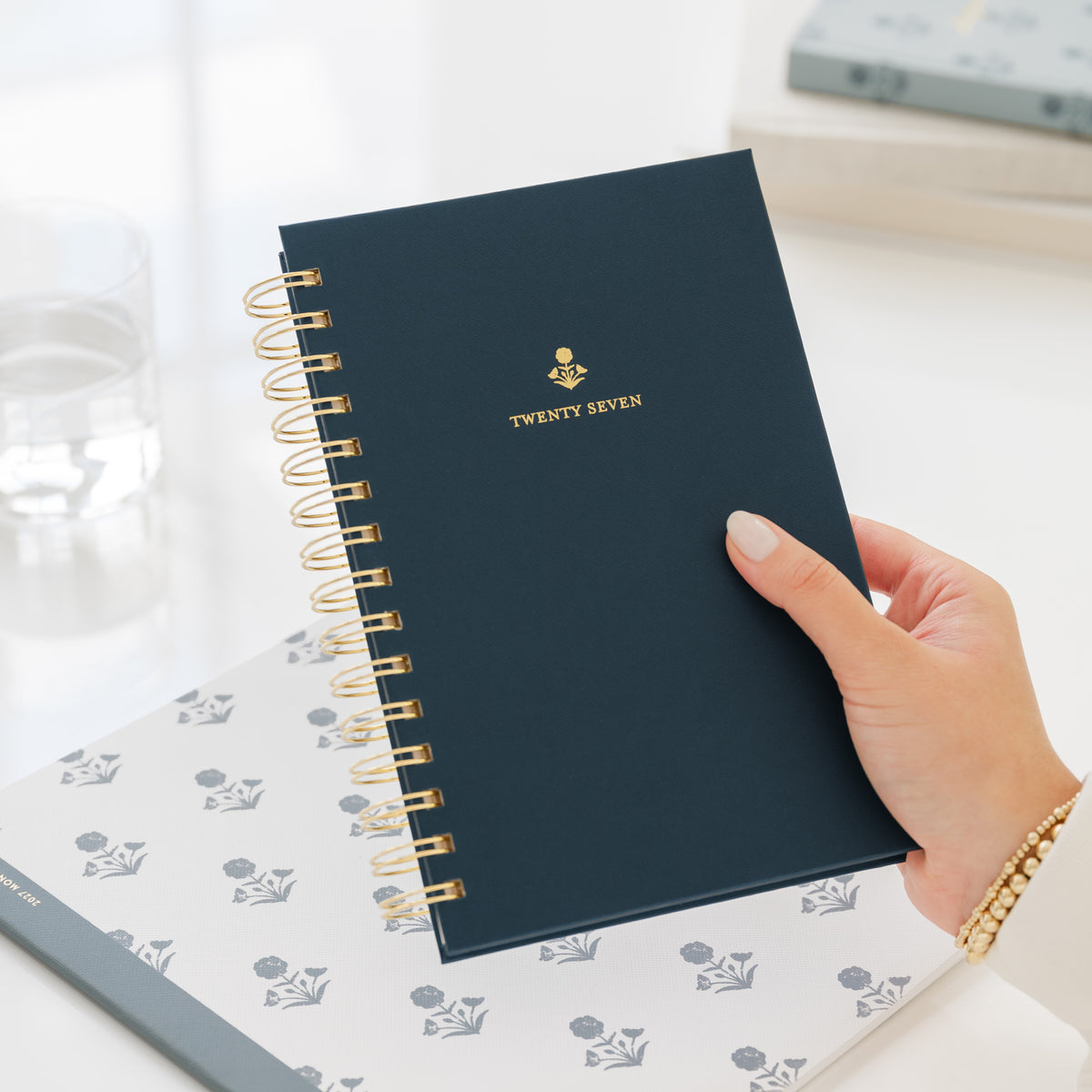 A hand holding a navy blue spiral-bound planner with gold wire binding and gold foil lettering that reads 'TWENTY SEVEN'. The planner is held over a white planner with a blue floral pattern