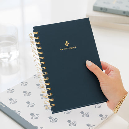 A hand holding a navy blue spiral-bound planner with gold wire binding and gold foil lettering that reads 'TWENTY SEVEN'. The planner is held over a white planner with a blue floral pattern