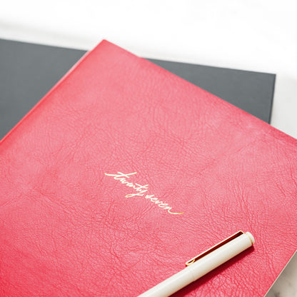 A red textured leather agenda with gold foil script reading 'twenty seven' on the cover, shown with a pale pink pen on a dark gray planner