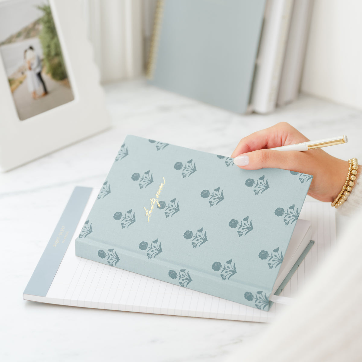 A light blue hardcover planner with a repeating floral pattern and gold foil script on the cover, held by a hand over an open lined legal pad