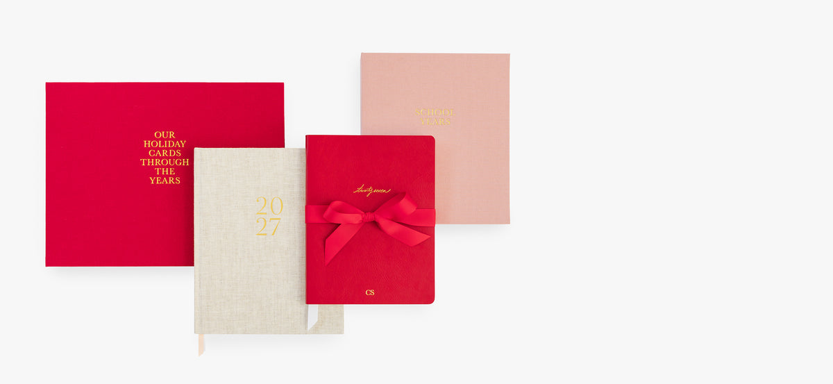 A red holiday card book, flax 2027 planner, red 2027 planner, and pink school years book (all with gold foil printing) on a white background.