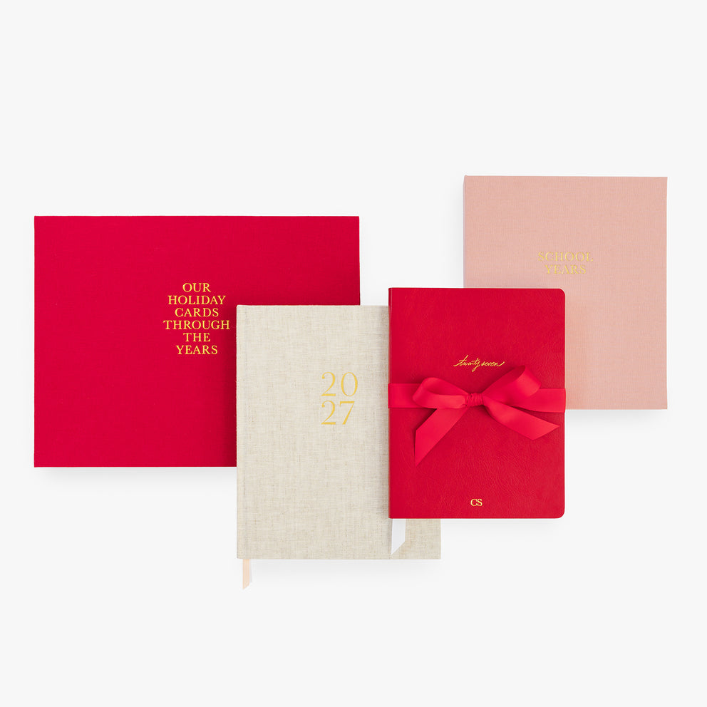 A coordinated set of books on a light gray background, including a bright red holiday cards book, a cream linen-textured 2027 journal, a red planner tied with a red ribbon, and a pale pink book with gold lettering.