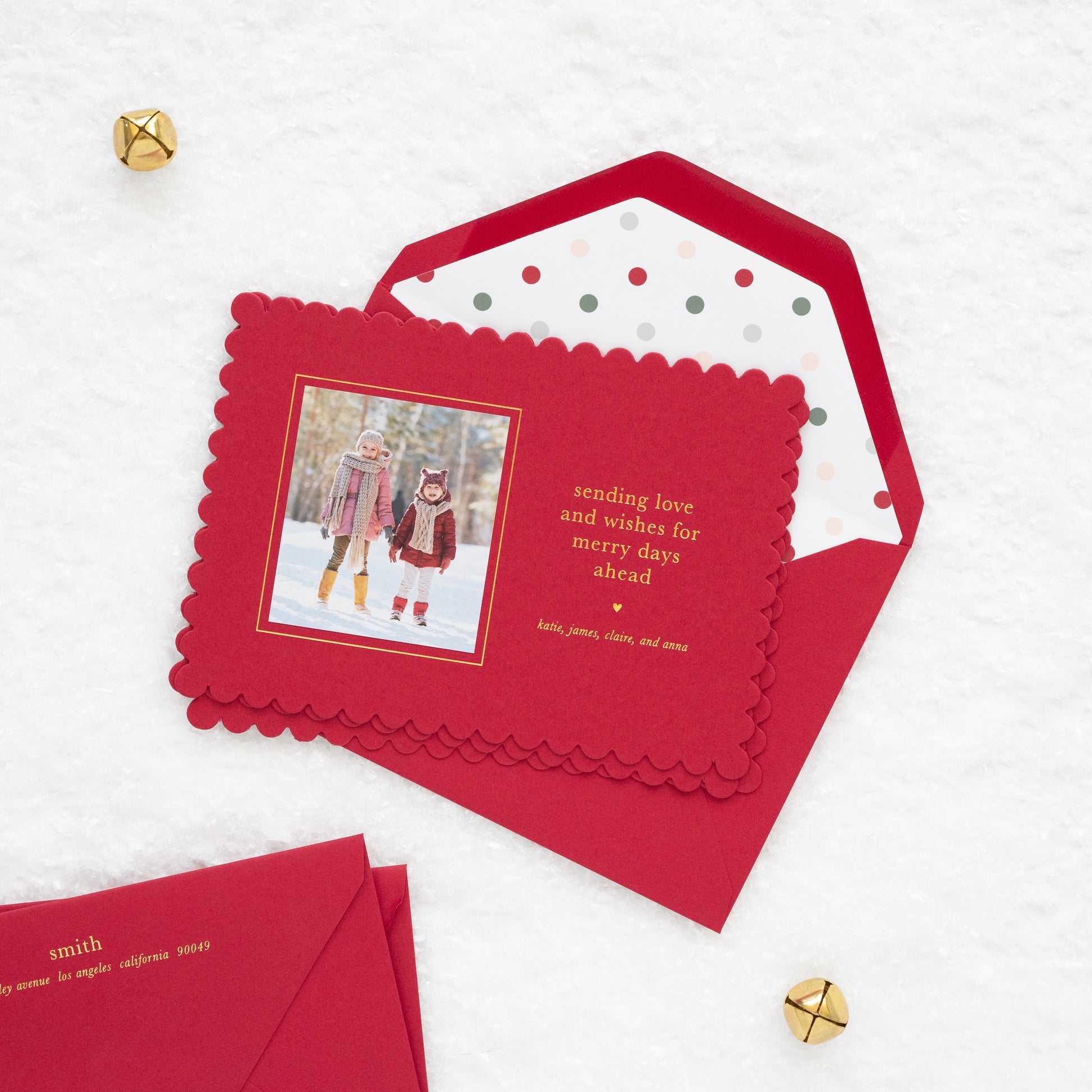 Close up of red scalloped custom holiday photo card with a photo of two kids in the snow with gold foil 'sending love and wishes for merry days ahead' paired with stack of red envelopes