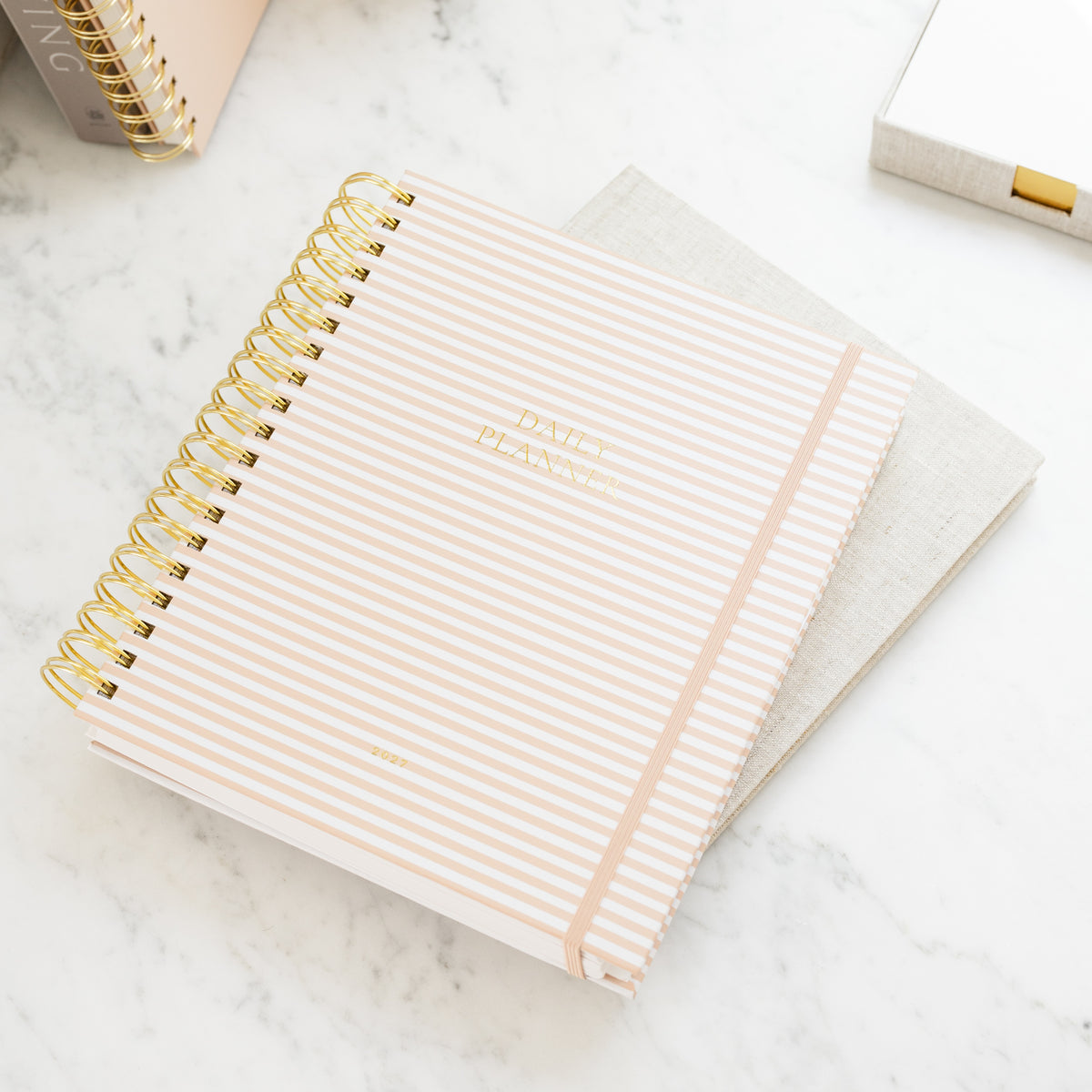 A striped daily planner with gold spiral binding and gold foil lettering on a marble surface