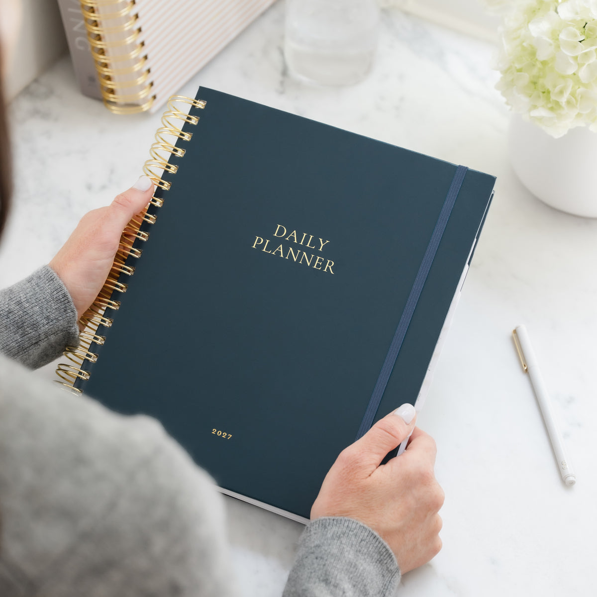 A navy blue spiral-bound daily planner with gold lettering printed with 'Daily Planner' and '2027' is held by a person wearing a gray sweater on a white marble desk. The planner features a dark blue elastic band and is shown alongside a white pen and a small vase of white flowers