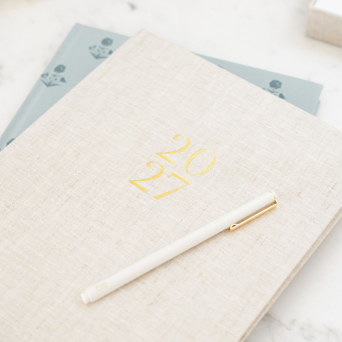 Flax linen fabric desk agenda printed with gold foil '2027' on the cover stacked on top of a blue floral desk agenda with a white pen on top