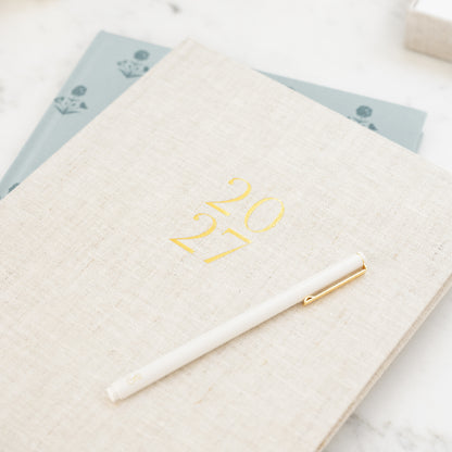 Flax linen fabric desk agenda printed with gold foil '2027' on the cover stacked on top of a blue floral desk agenda with a white pen on top
