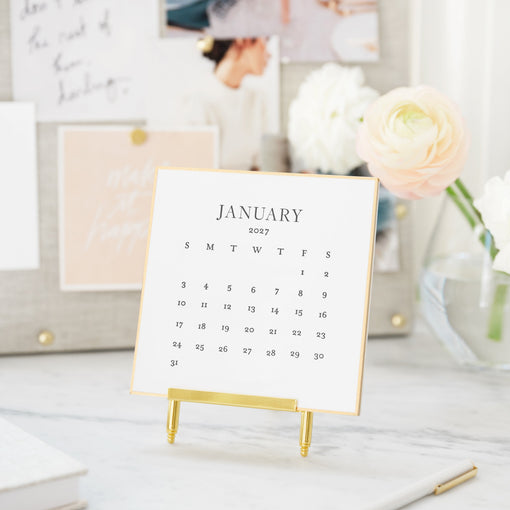 A small January 2027 desk calendar with a gold frame and gold stand, displayed on a marble desk in a styled home office setting with flowers and pinned notes in the background.