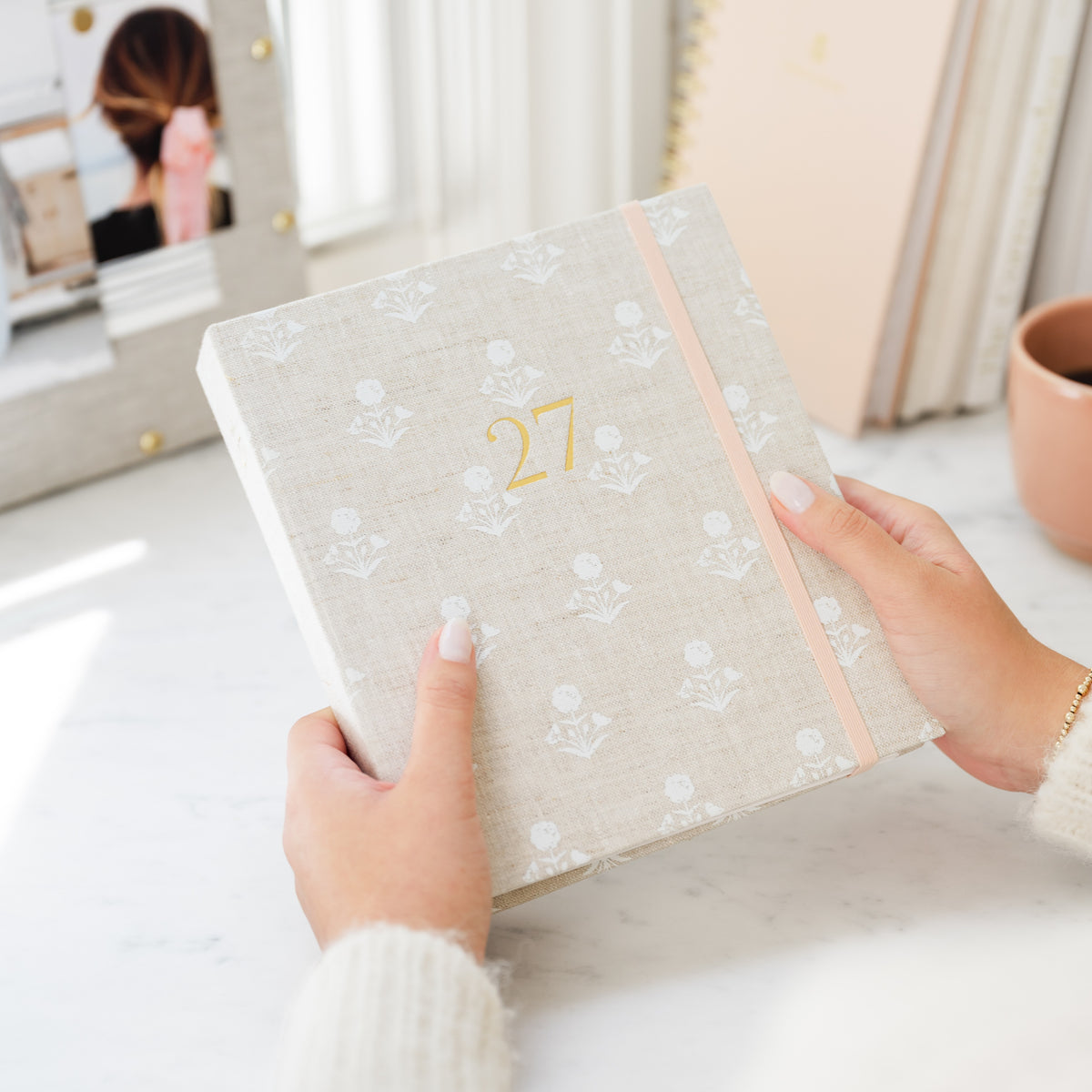A person holds a linen-covered planner with a pale pink elastic band and the number 27 in gold on the cover. The setting is a bright, modern room with a marble surface, and an inspiration board and cup of coffee nearby