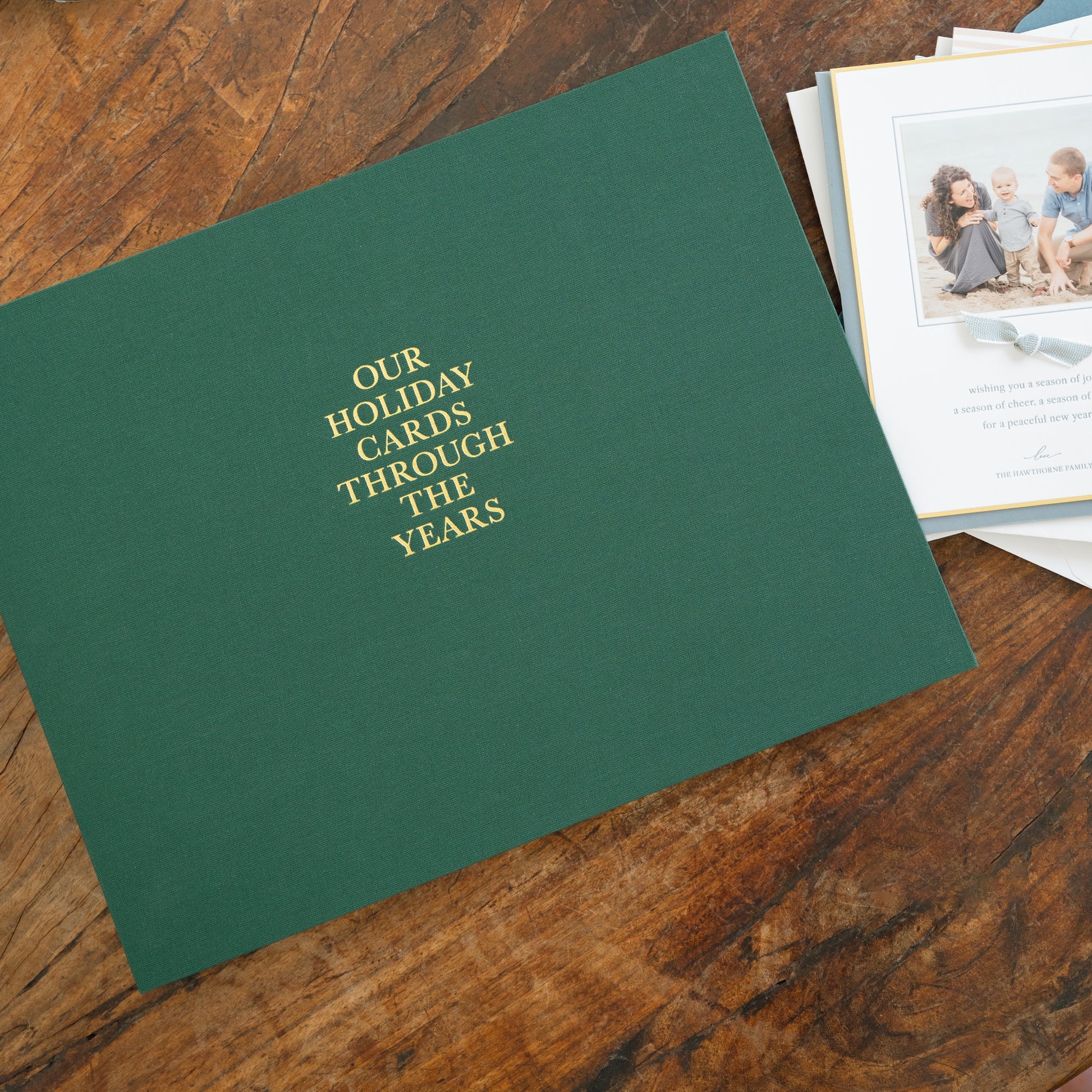 Dark evergreen holiday card book on wood table with family holiday photo cards on side