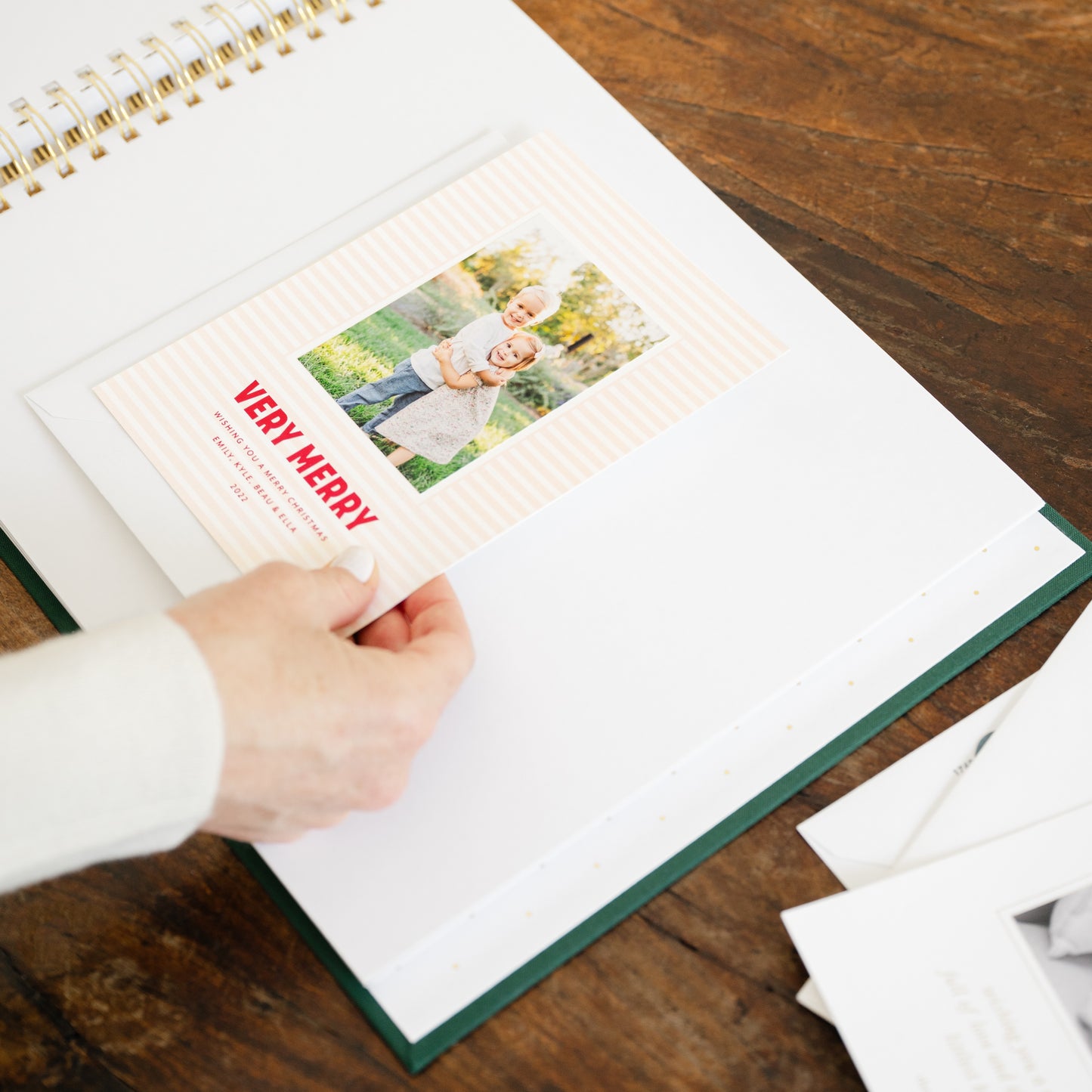 A person's hand inserting a printed holiday card into a white spiral-bound album with a dark green cover, placed on a wooden table.
