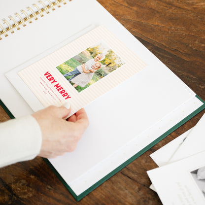 A person's hand inserting a printed holiday card into a white spiral-bound album with a dark green cover, placed on a wooden table.