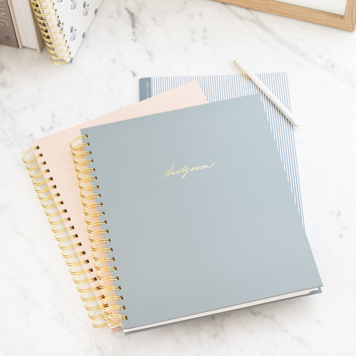 A stack of spiral-bound planners on a white marble surface. The top notebook is dusty blue-gray with gold foil script reading 'twenty-seven' and gold spiral binding.