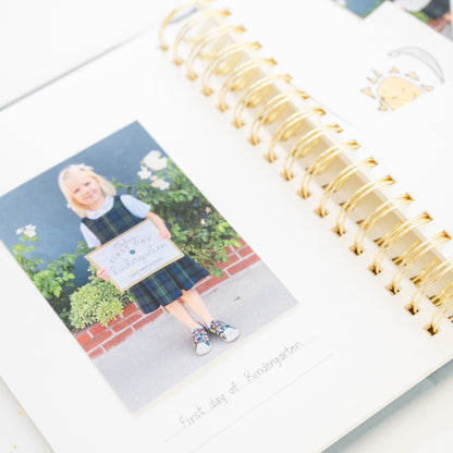 A close-up of a spiral-bound photo album with gold rings, showing printed photographs of a child on white page in their school uniform written beneath is "first day of kindergarten"