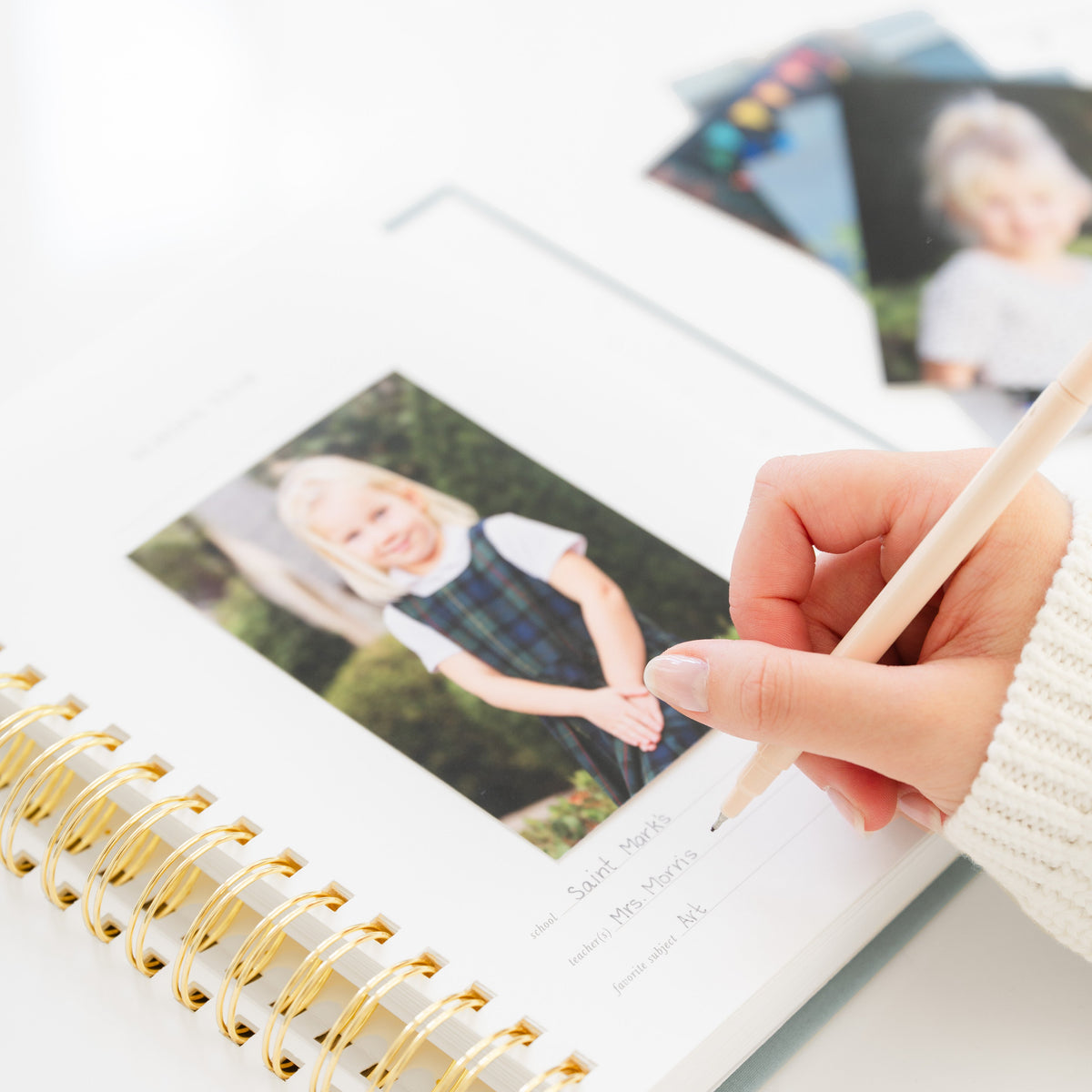 A spiral-bound photo album with gold rings on a white surface, showing printed children's school photos and a hand holding a pencil pointing at one page.