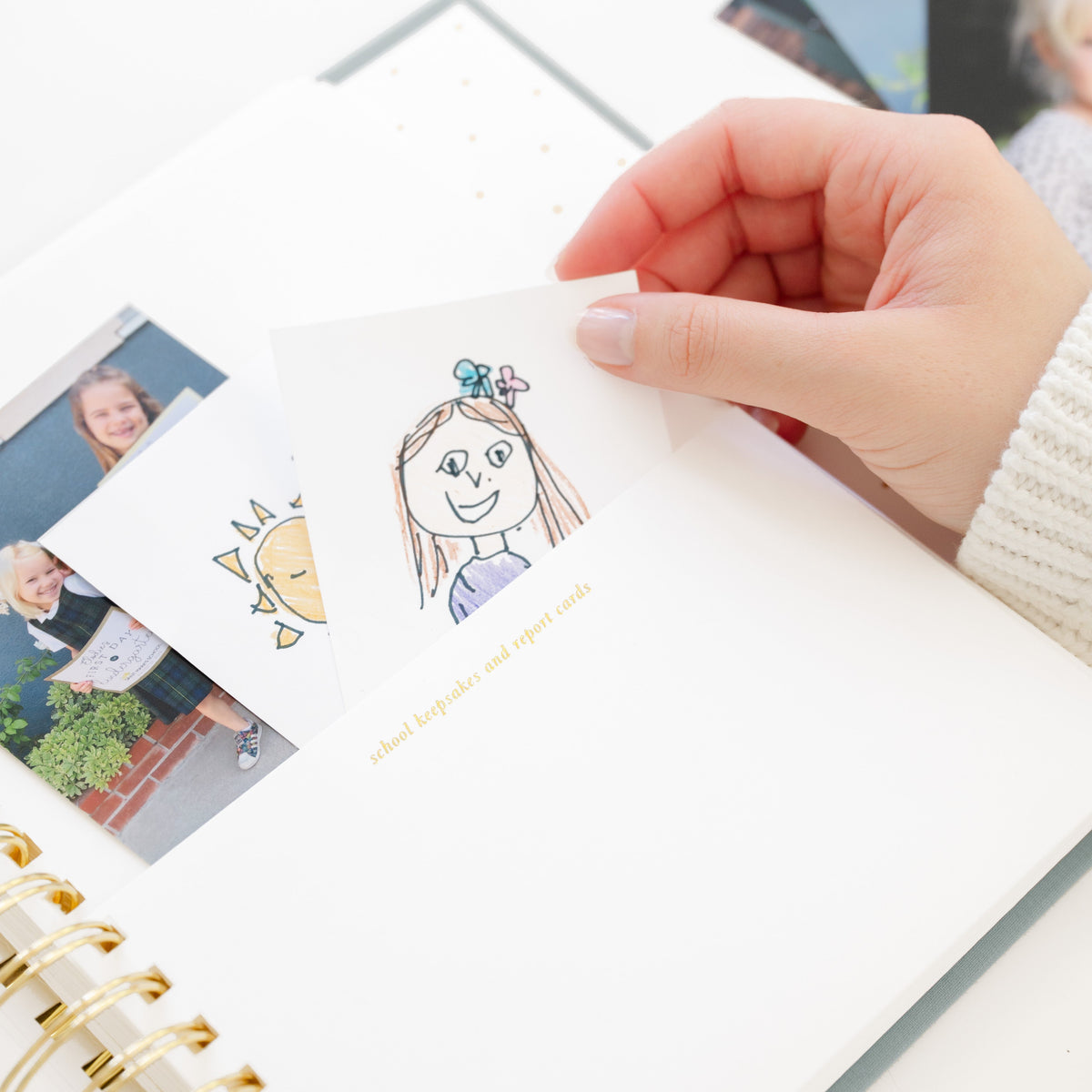 A hand holding a small hand drawn card with a cartoon girl illustration going into a pocket with gold foil printing 'school keepsakes and report cards'