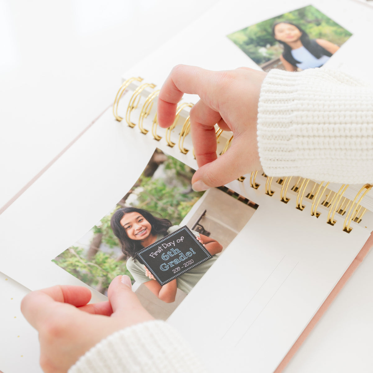 A close-up of hands turning pages in a spiral-bound photo album with gold rings, showing printed photos of a woman outdoors on a bright white surface.