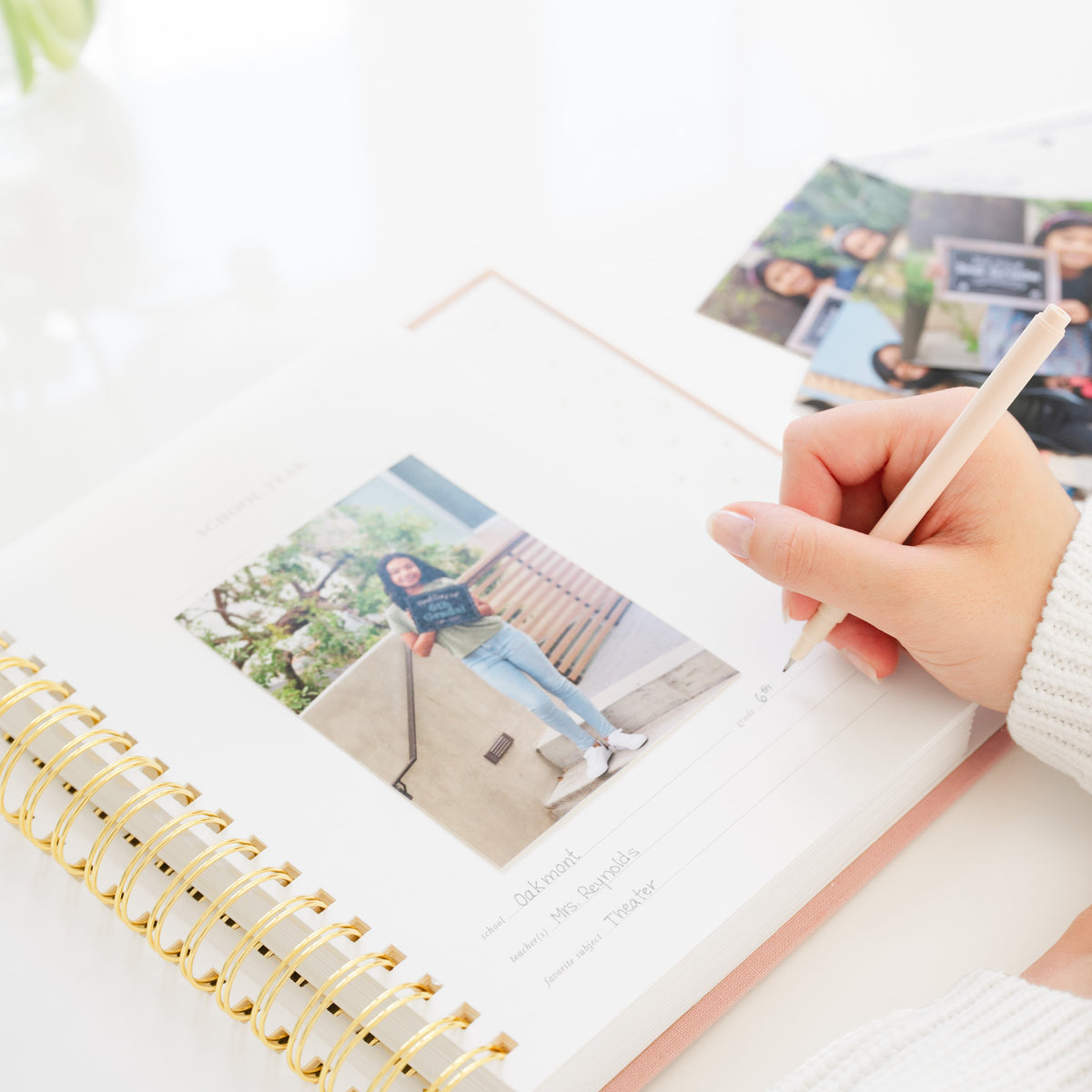 A hand holding a pencil writes in a spiral-bound photo keepsake school years book with a gold spiral, with a line for "school," "teacher," and "favorite subject" with printed photos in background