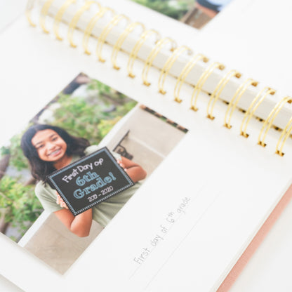 A spiral-bound photo album or planner with gold wire binding, shown open on a white surface. The pages contain printed photos of a young girl holiding a sign that says 'First Day of 6th Grade!' and 'first day of 6th grade' written on a line below.