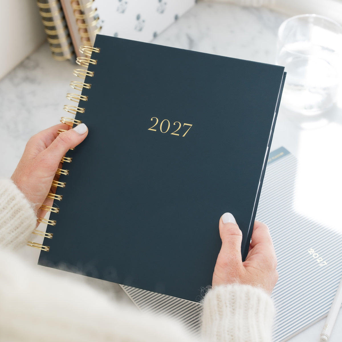 A person holding a navy blue 2027 spiral-bound planner with gold wire binding and gold '2027' text on the cover over a kitchen counter with other planners and a glass of water in background