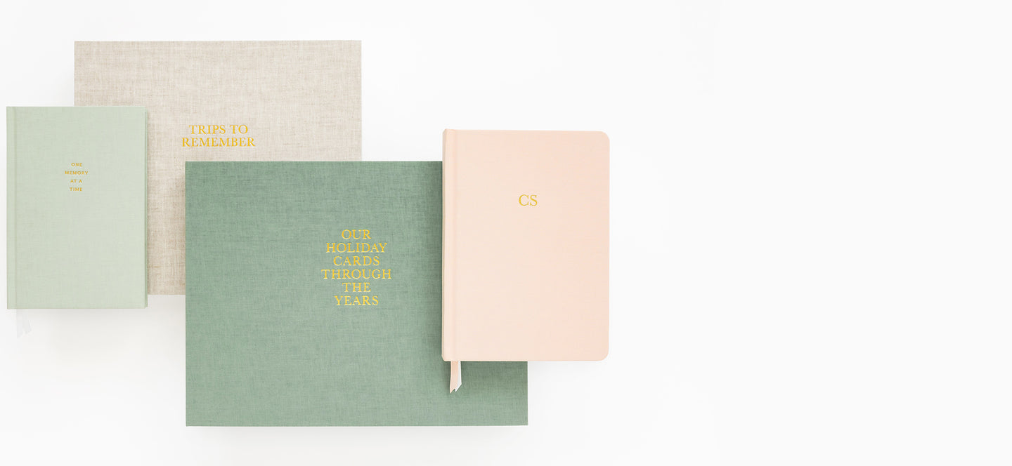 stack of fabric keepsake books in flax, green and pink including one memory at a time journal, trips to remember, our holiday cards through the years, and a pink journal with initials foil stamped on the cover