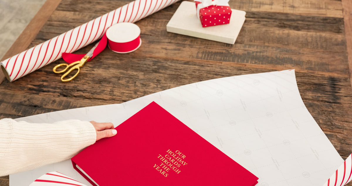 A person's hand in a white sweater holds a red gift box wrapped in white paper printed with 'Our holiday cards through the years' in gold foil. The scene is set on a rustic wooden table with holiday-themed wrapping supplies, including a roll of red and white striped paper, a spool of red ribbon, and gold scissors.