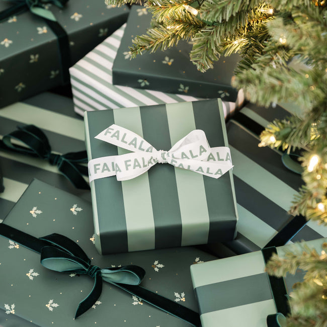 A stack of wrapped Christmas gift boxes in dark green, sage green, and white striped paper, tied with black ribbons and a white ribbon printed with the brand name 'FALALA'. The scene is styled under a lit Christmas tree branch.