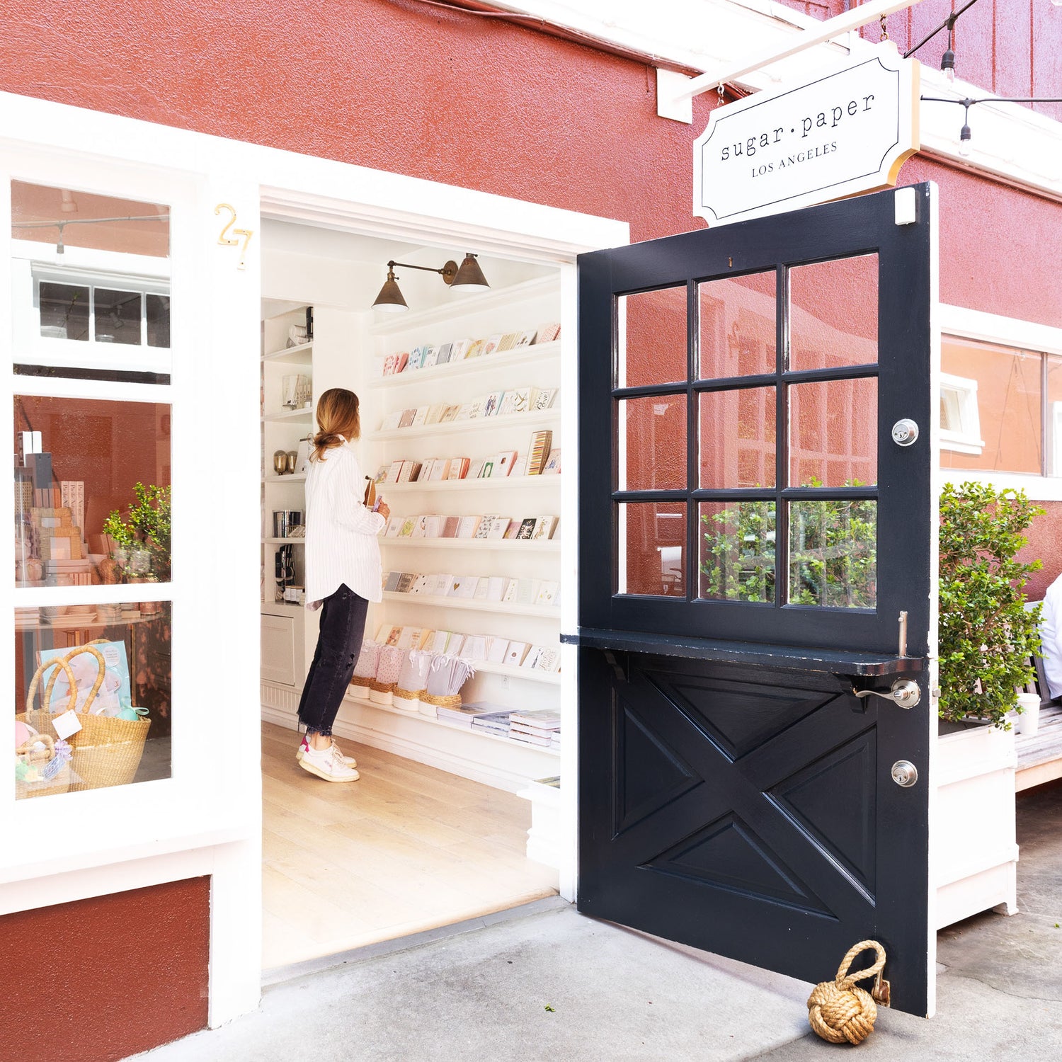 A storefront scene with a person standing inside a book-filled shop next to an open black door with a sign reading sugar paper LOS ANGELES.