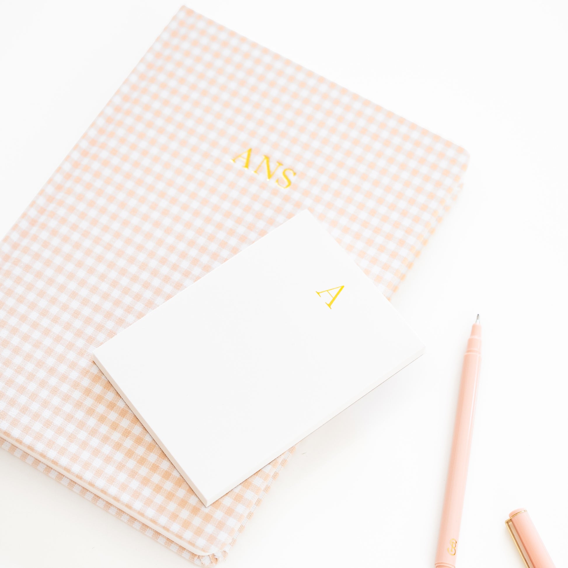Pink gingham fabric journal with 'ANS' on cover paired with a white mini notepad with 'a' in gold foil and a pink pen