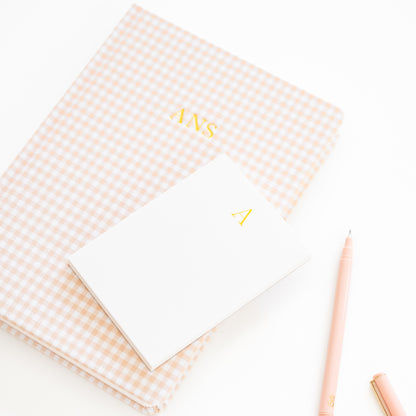 Pink gingham fabric journal with 'ANS' on cover paired with a white mini notepad with 'a' in gold foil and a pink pen
