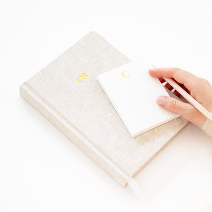 Hand holding a white notepad with gold 'c' on top of a flax journal with 'cs' lettering in gold foil