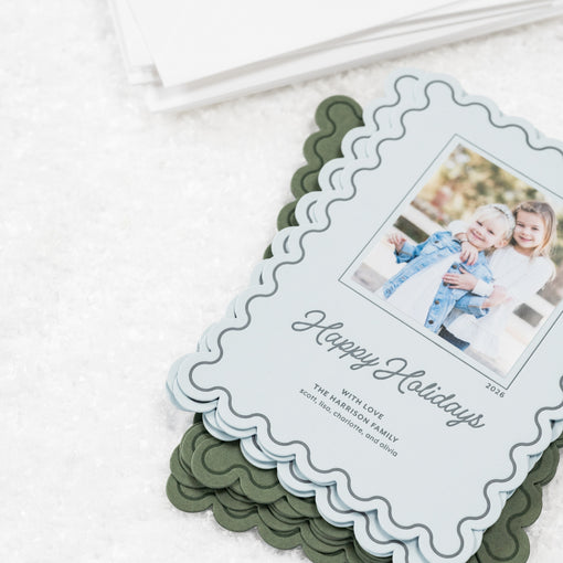 A close-up of a stack of scalloped light blue holiday photo cards on top of a stack of green scalloped holiday photo cards placed on a white textured surface with snow. The card includes a printed photo of two children and the text 'Happy Holidays' with personalized details below.
