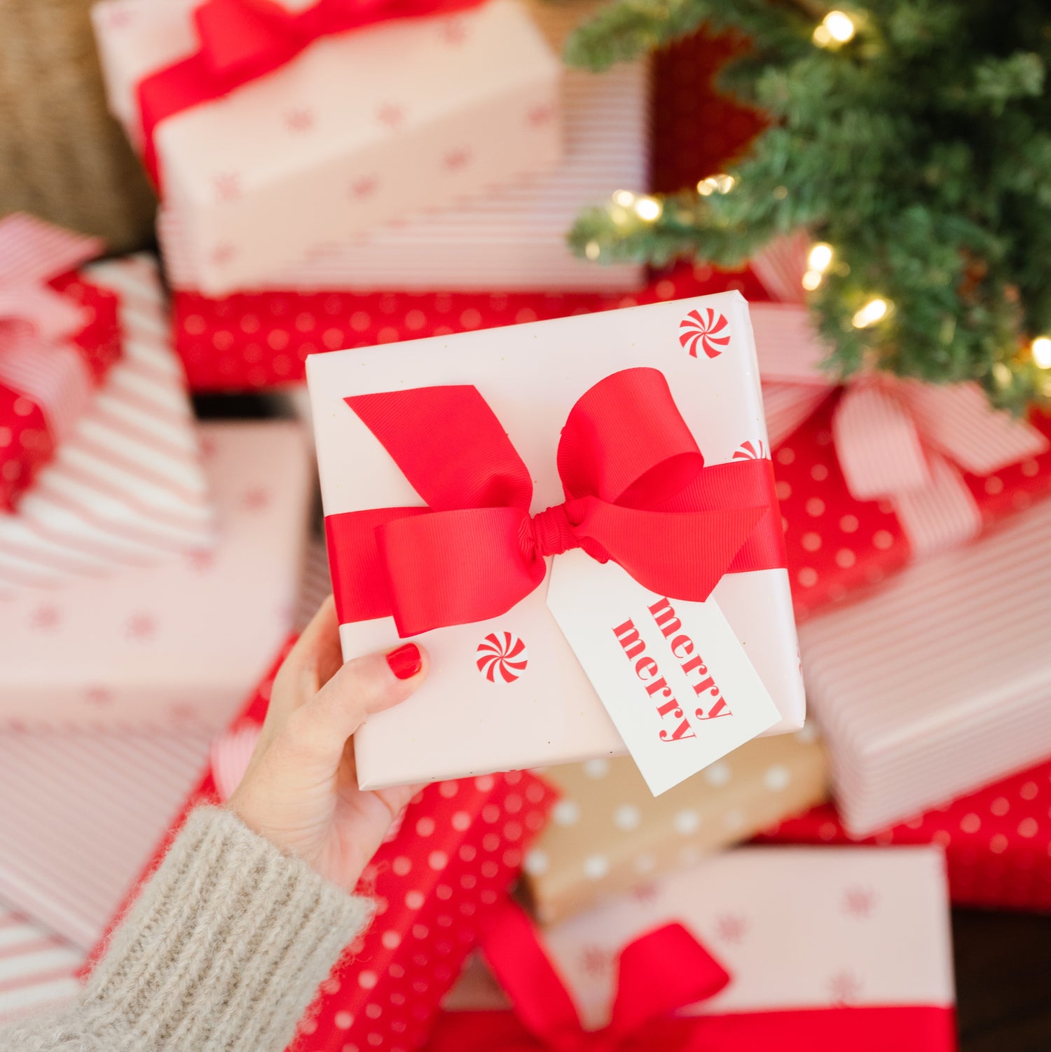 Hand holding a Christmas gift with a red bow and 'Merry' tag, surrounded by other wrapped gifts.