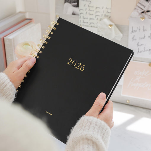 Black spiral planner with '2026' on the cover held in hands at a desk setting
