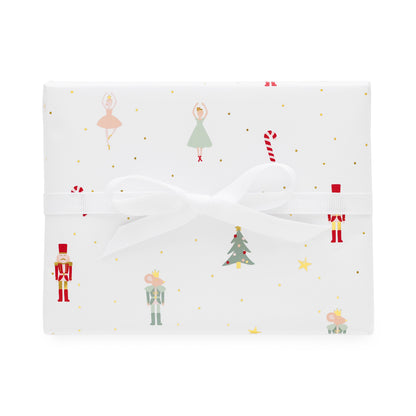 A square gift box wrapped in white wrapping paper with a festive Christmas print featuring nutcrackers, ballerinas, candy canes, a Christmas tree, and gold dots, tied with a white ribbon