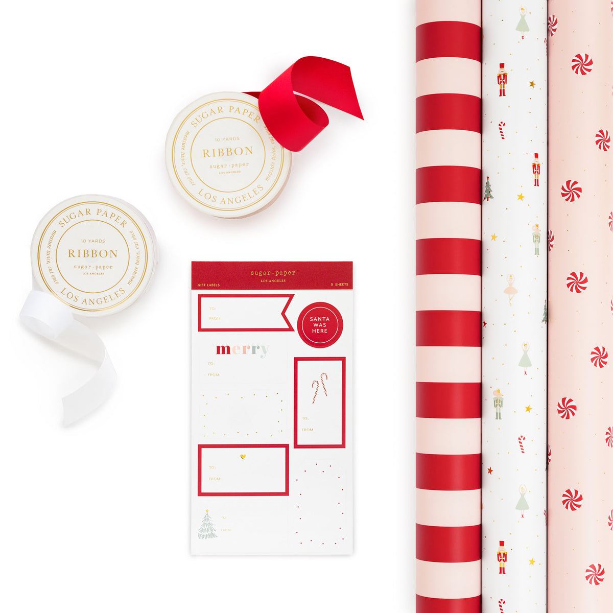 A flat-lay of holiday wrapping supplies on a white background, including two rolls of Los Angeles Sugar Paper ribbon with gold labels and a red ribbon bow, a sheet of holiday stickers, and three rolls of wrapping paper with red-and-white stripes, white with red candy canes, and white with red peppermint candies.