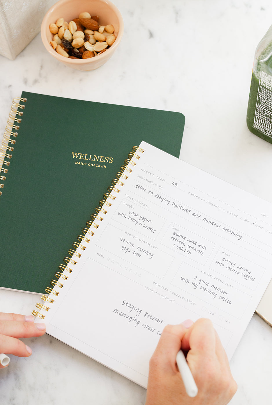 Person using a wellness daily planner with a green cover on a marble surface.