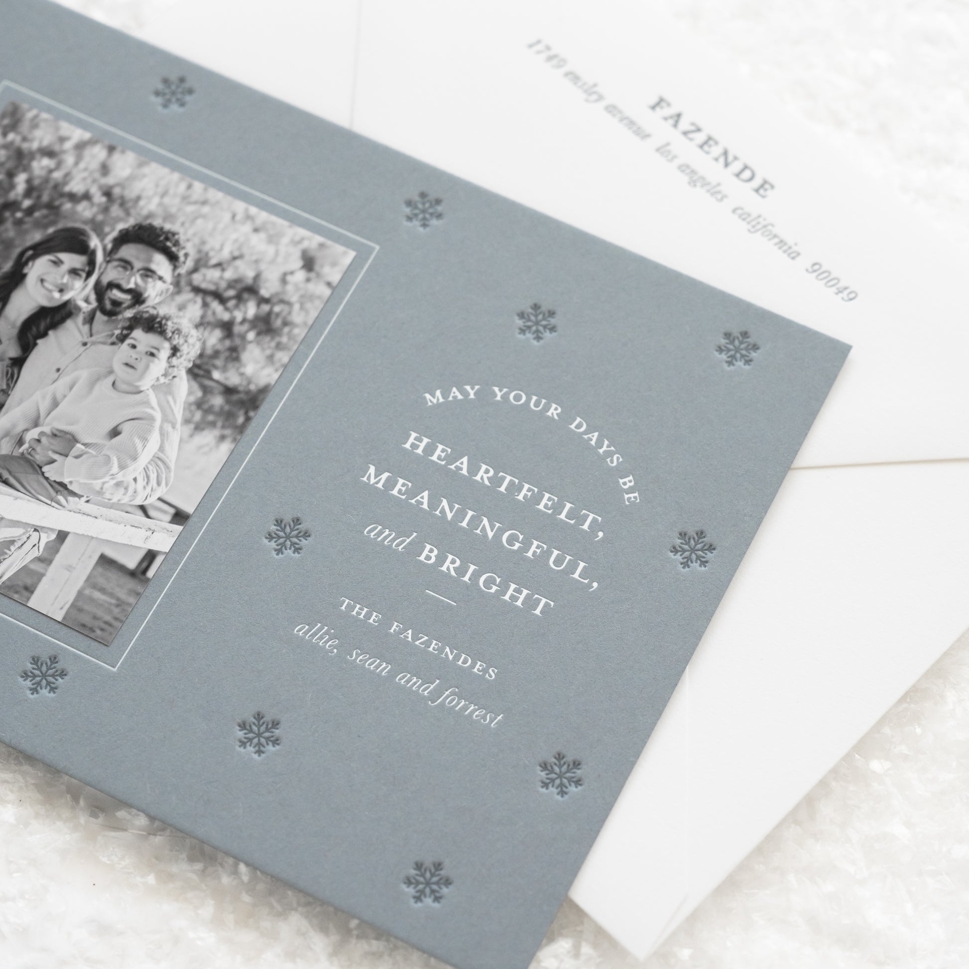 Close up of coastal blue custom holiday photo card paired with a black and white photo with white foil greeting 'may your days be heartfelt, meaningful, and bright' with dark blue snowflake pattern letterpress printed paired with a white envelope